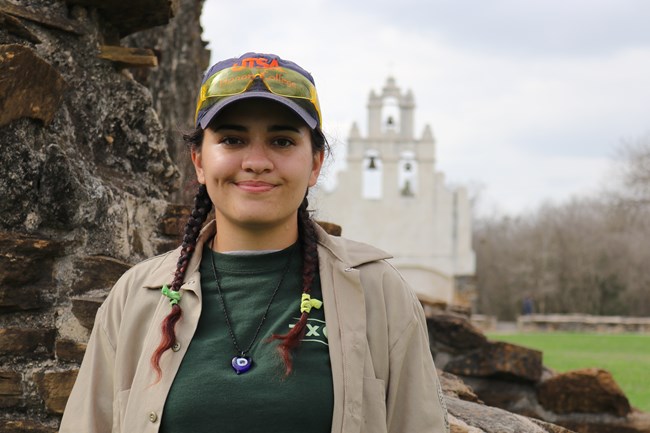Female apprentice, a young adult, smiles in foreground with Mission San Juan church facade in background.