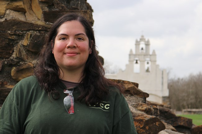 Sarah, a young woman wearing green, stands in front of the limestone church facade at Mission San Juan.