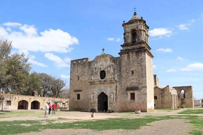 Limestone church facade at Mission San Jose midday, with scattered visitors on surrounding pathway.