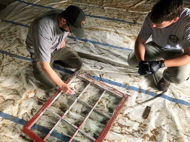 Two NPS employees in uniform work on an old window. The window frame is painted red but the paint is peeling.