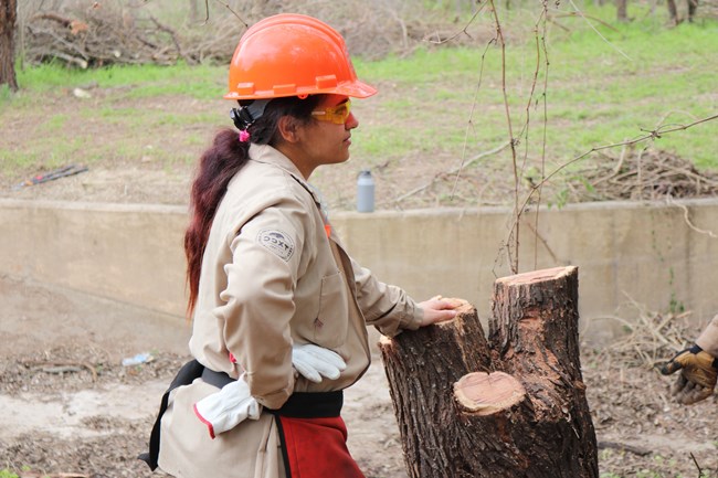 Karli leans on a tree stump while wearing a bright orange hard hat