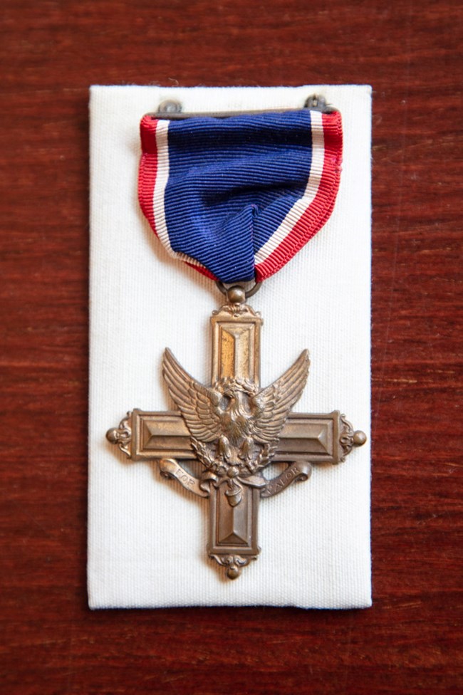 Metal cross with eagle emblazed on front held by red, white, and blue cloth.