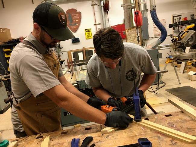 Two people use clamps on a wooden frame in a workshop. One of the people is wearing an NPS uniform while the other is in an intern uniform.