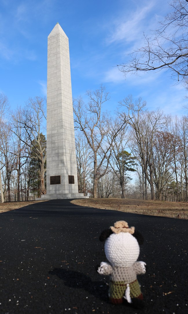 Ranger Cow stands on paved path looking toward a tall monument