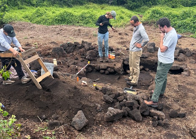 Two male and one female archeologist stand looking at partially excavated pit with stacked stones