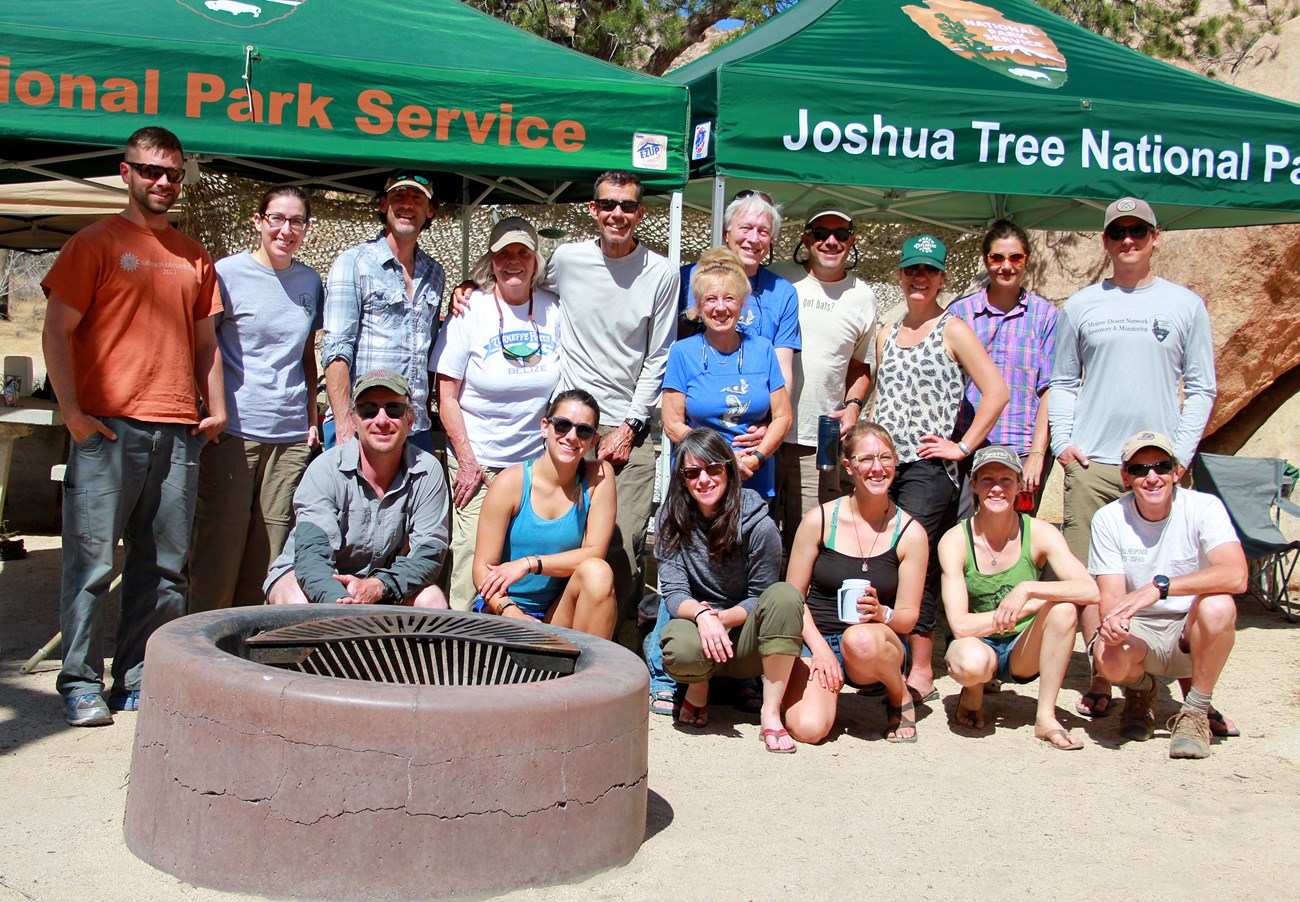 A large group of people sitting or standing in front of two covered shelters.