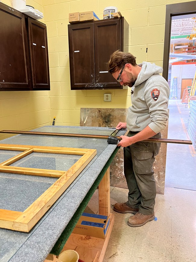 HPTC staff at FOVA In the foreground is a wooden window, with all the paint stripped off, laying on a large table. In the background, a man in a National Park Service uniform checks measurements on another piece of wood.