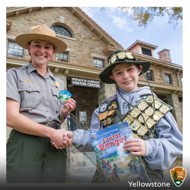 A junior ranger with many badges shakes the hand of a park ranger.