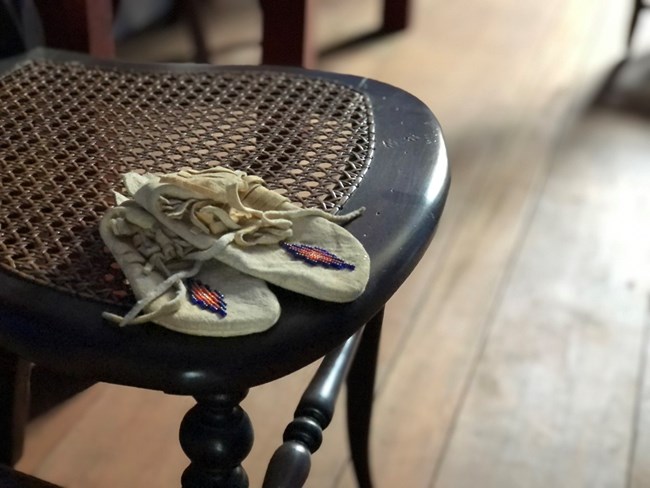 A pair of small, child-size beaded moccasins sitting on a wooden chair with a caned seat.