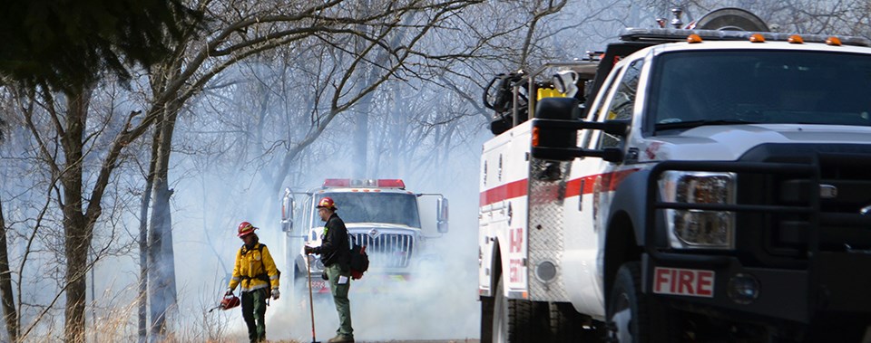 Cowles Dune Prescribed Fire - 2017 A firefighter uses a drip torch to ignite vegetation while standing near engines and another firefighter.