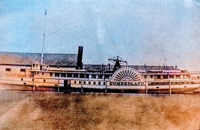 SS Cumberland docked with large building behind it, sidewheel displaying name in large letters