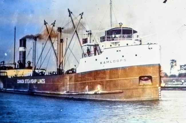 view of the port side of the KAMLOOPS under power with cityscape in distant background and small boat directly behind