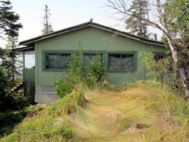 green cabin with four prominent windows in front, long grass growing in the foreground, water in the distance