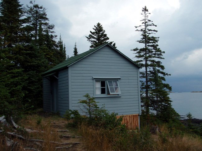 small cabin with blue wood siding and an open window, water in the background