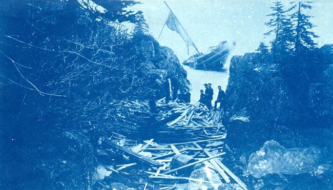 barrels and boards from SS Algoma are strewn between a gap in the outer rocks of Mott Island, ship sinking in the background, people looking on