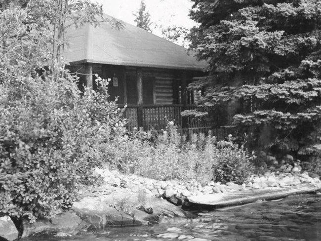 square cottage somewhat hidden behind dense brush, open covered porch, triangular roof