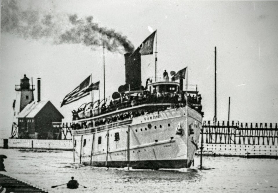 SS Puritan sailing out of St. Joseph Harbor, decks swarmed by passengers, onlookers in rowboat