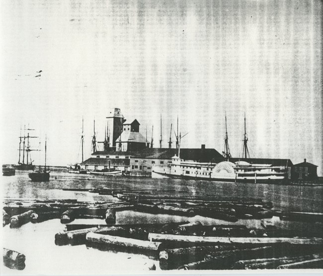 SS Cumberland docked with large building behind it, logs in harbor in foreground
