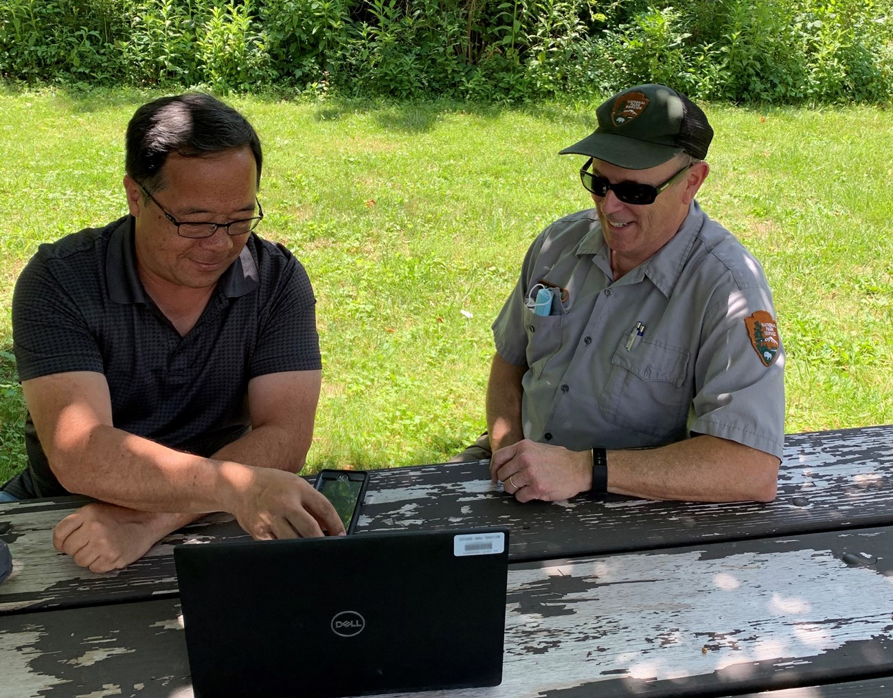 Two men sit outside at a picnic table; the man to the left wears a gray polo shirt and types on a black laptop; the man on the right wears a gray uniform shirt and green hat, which both contain the brown, green and white NPS arrowhead symbol.