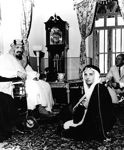 King Ibn Saud seated in a wheelchair in a room with other men.