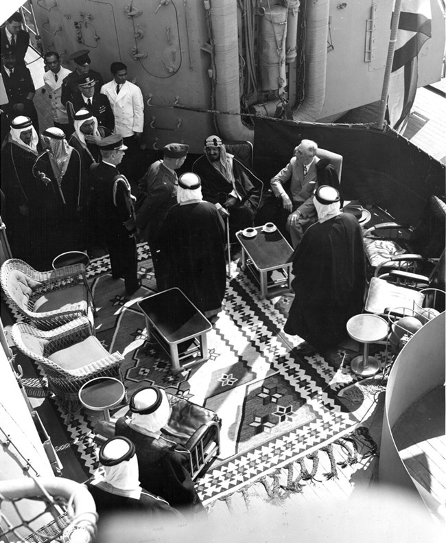 A group of men surrounding Ibn Saud and FDR seated on the deck of the USS Quincy