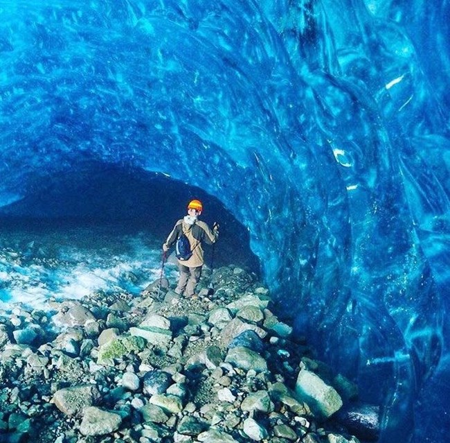 Ice Cave Root Glacier WRST with person a person wearing a climbing helmet walking into an ice cave on a steep rocky slope
