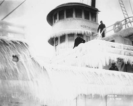 IceShip Two sailors, dressed in dark clothes, stand on the desk of an ice encrusted ship. The words “North Lake” are painted on the side of the ship. The ice covering the boat is so thick in spots that the shape of the boat itself is obscured.
