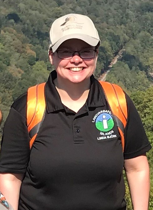woman with glasses and a polo shirt smiling at the camera with a mountain in the background