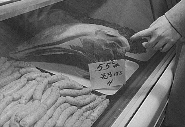 Black and white photo of a grocer’s meat case showing a cut of beef and sausages. The sign in front of the beef reads 55c per pound, 8(crossed out) 4 points. A white hand is gesturing at the meat as though selecting it.