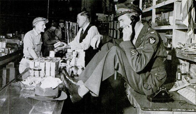 Black and white photo. Stewart, in full uniform, sits on a shop counter and talks on the phone. His father is in the background, helping a customer. The shelves are full of goods.