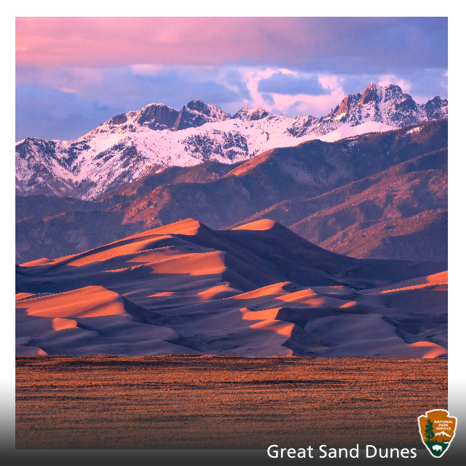 A warm sunset glow illuminates sand dunes and snowy mountains.