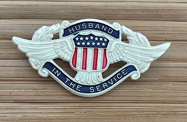 White plastic pin. In the center is a shield with red, white, and blue stars and stripes. Eagle wings stretch out from the sides, framed by a laurel wreath. The banners with the text are blue.