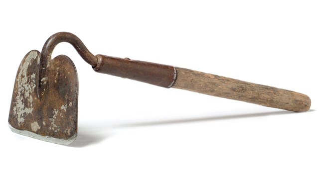Color photo of a short-handled hoe. It has a wooden handle and a rusted iron blade.