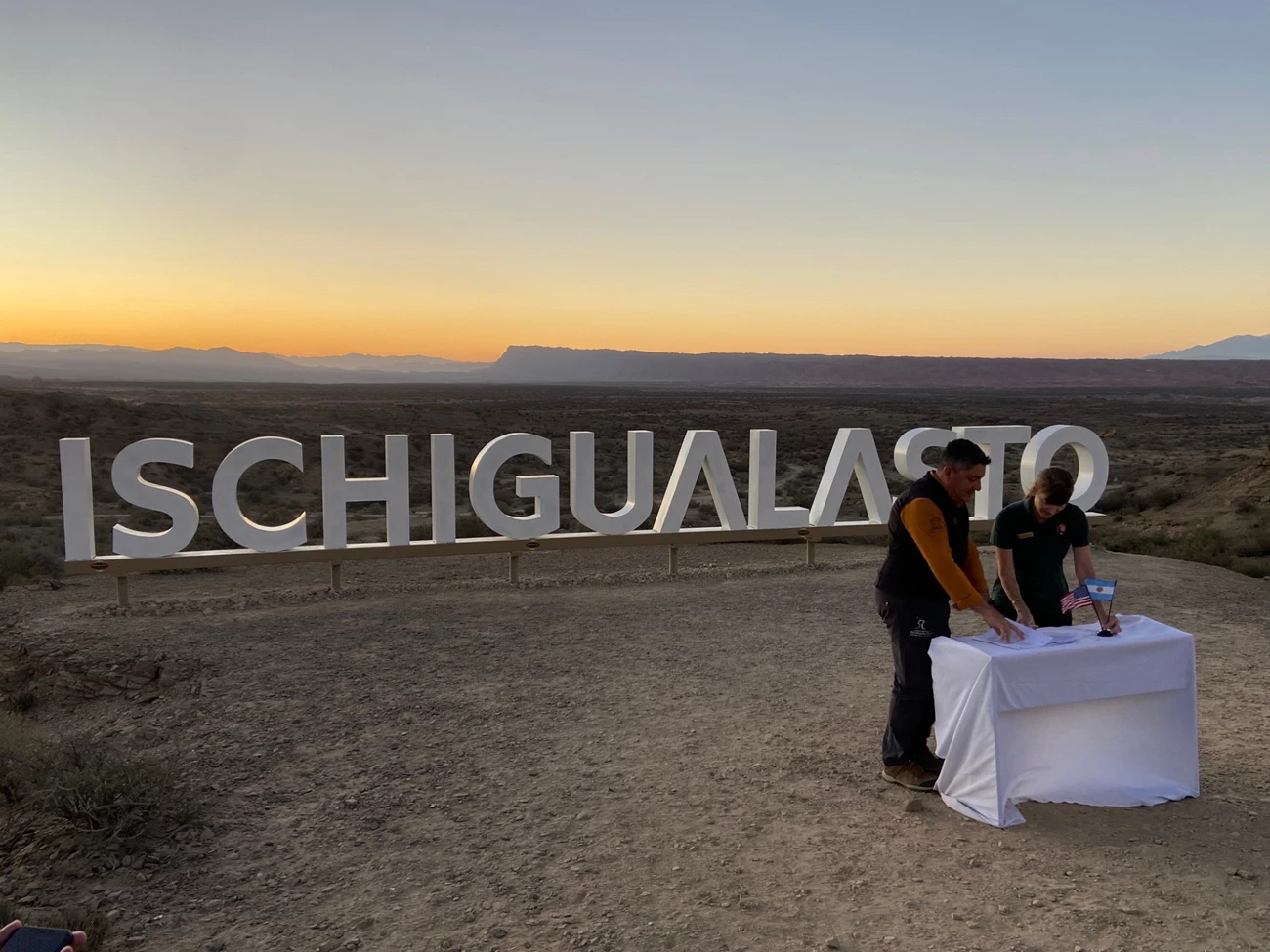 Image C Photo two people at a small table in front of a sign for Ischigualasto.