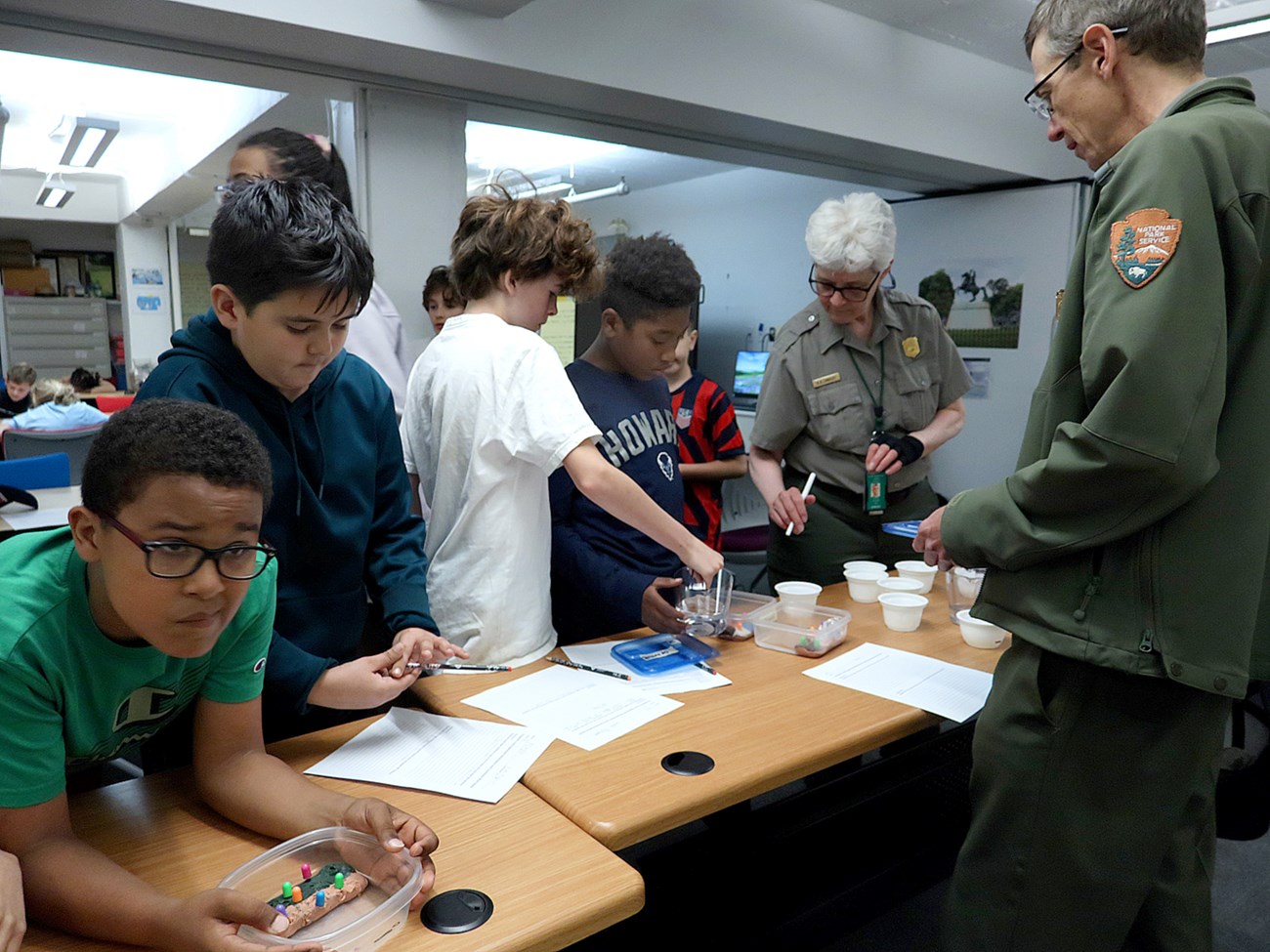 A group of children stand around a counter holding containers. A man and woman in NPS uniforms look on.