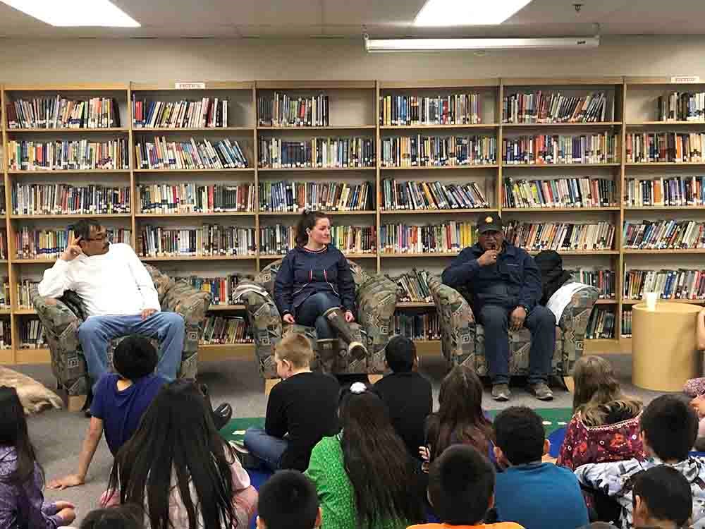 Image_library_web Three speakers sit in a library addressing a group seated on the floor in front of them.