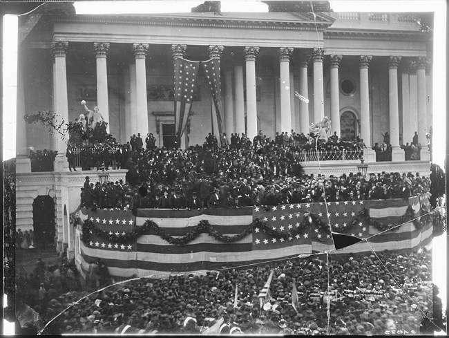 Black and white image of the White House with people lined on the balcony and below, and flags covering the white house