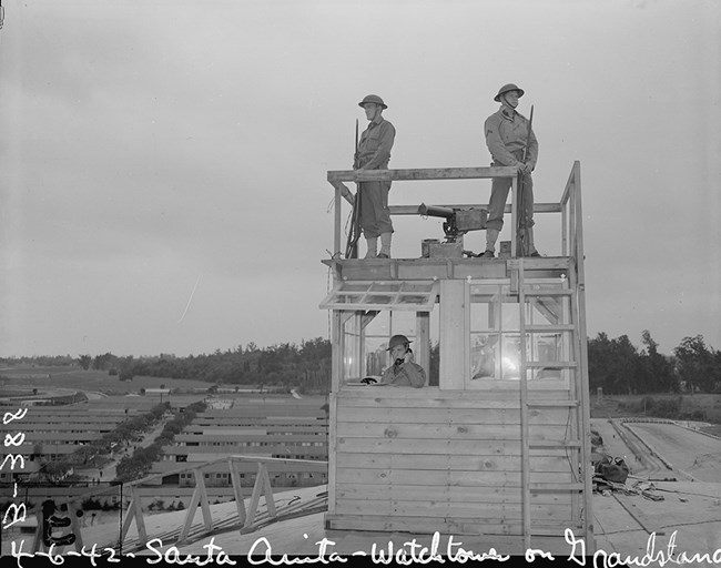 Black and white photo. Two guards armed with rifles stand watch on a wooden guard tower. The guns have bayonets. Inside the tower is another guard speaking into a telephone. The racetrack and horse stalls are in the background.