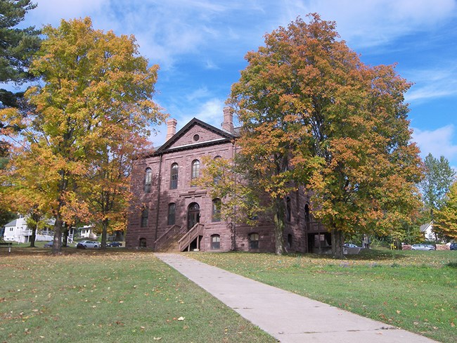 Color photo. A three-story building of red sandstone sits between two trees just beginning to turn colors in the fall. An area of grass and a sidewalk lead up to the building. The sky is bright blue.