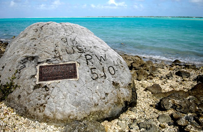 Color photo. The large coral rock sits on a rocky beach at the edge of a turquoise-colored lagoon. The grey stone also has a weathered brass commemorative plaque.