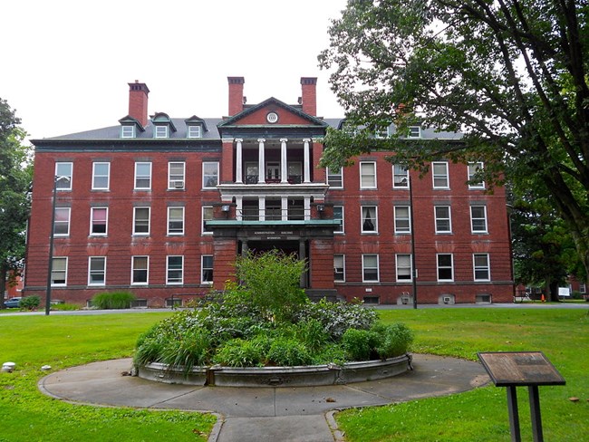 Color photo. A large 5-story red brick building with white columns and a sign reading “Administration Building” over the main door. A concrete path leads across the lawn to the entrance, traveling around a raised ornamental garden bed. The sky is grey.