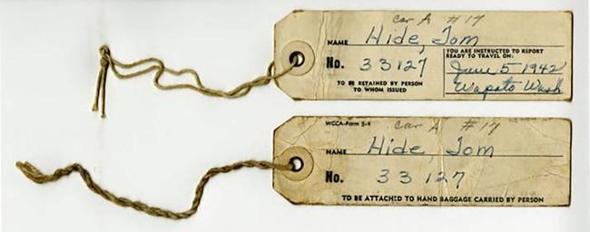 Two tan-colored paper tags on strings. Details are written in blue ink. They indicate that the Hide family was to report to Wapato, Washington on June 5, 1942. A pencil notation indicates they traveled in Car A, #17.