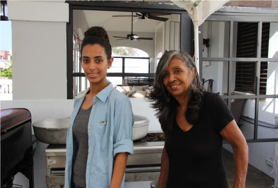 Indra Brugueras (left) and her abuela (grandmother) Isabel Pizarro work in their makeshift kitchen. Two women stand smiling in a makeshift kitchen.