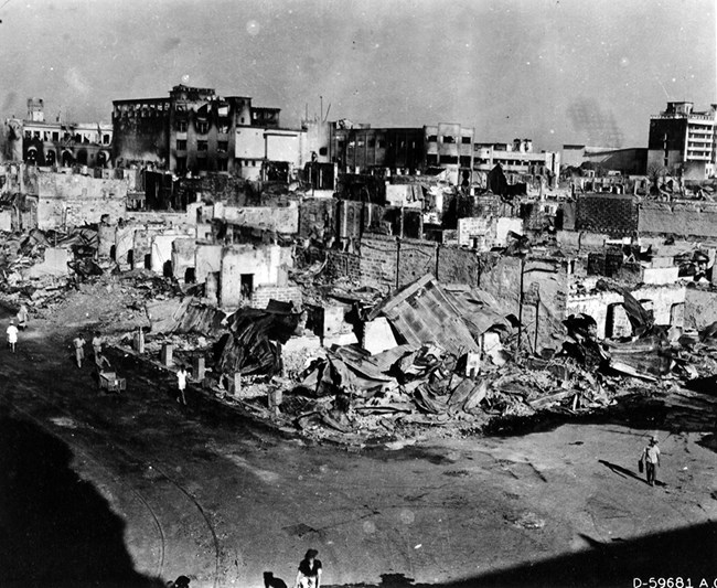 Black and white photo overlooking ruins of buildings. In the foreground, only a few walls are standing. In the distance, windows are blown out and there is evidence of fire. A handful of people wander the streets.