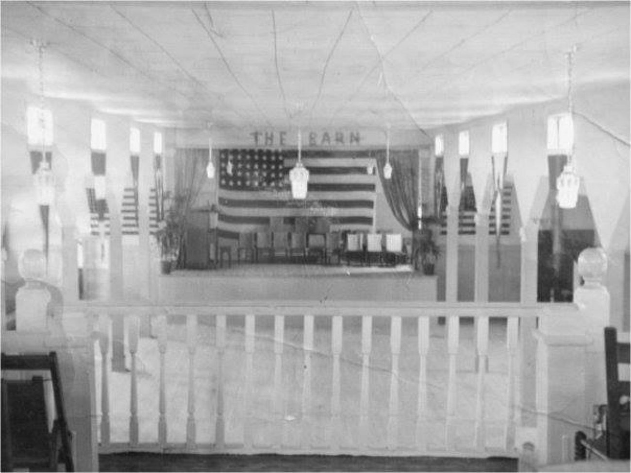 A stage room with lanterns hanging from the ceiling and an American flag hung on the back wall. There is a fence separating the room from another that is not pictured. "The Barn" is written on top of where the flag hangs