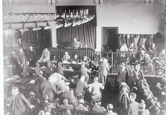 A large group of men are standing and seated in the interior of a large courtroom with wooden chairs and a chandelier hanging from the center of the ceiling. A judge sits in the center.