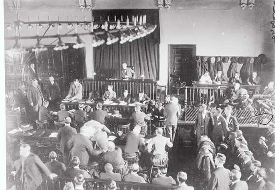 Interior Courtroom A large group of men are standing and seated in the interior of a large courtroom with wooden chairs and a chandelier hanging from the center of the ceiling. A judge sits in the center.