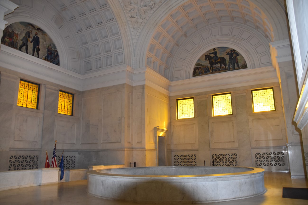 Photo of the interior of a mausoleum, featuring domed ceilings, white walls with architectural detailing, and a marble floor