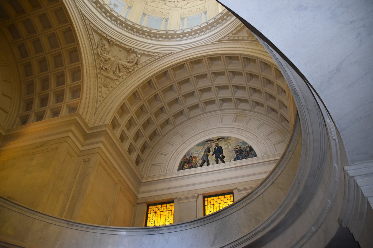 Photo of the interior of a mausoleum, featuring domed ceilings, white walls with architectural detailing, and a marble floor