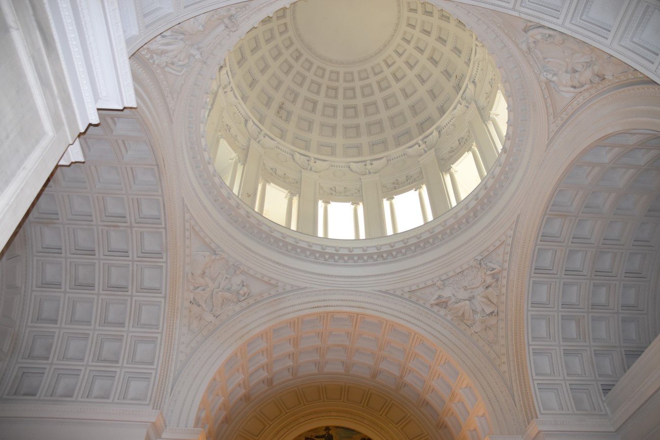 Photo of the interior of a mausoleum, featuring domed ceilings, white walls with architectural detailing, and a marble floor